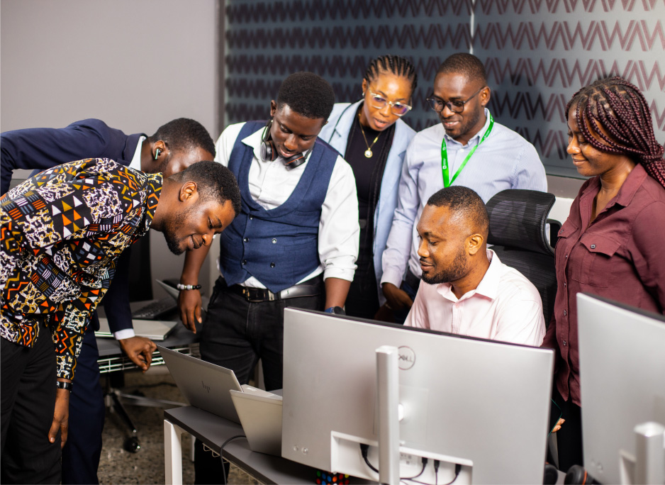 Seven individuals are gathered around a computer desk, concentrating on the screen of a desktop computer. One person is pointing at the screen, explaining or highlighting something to the group.