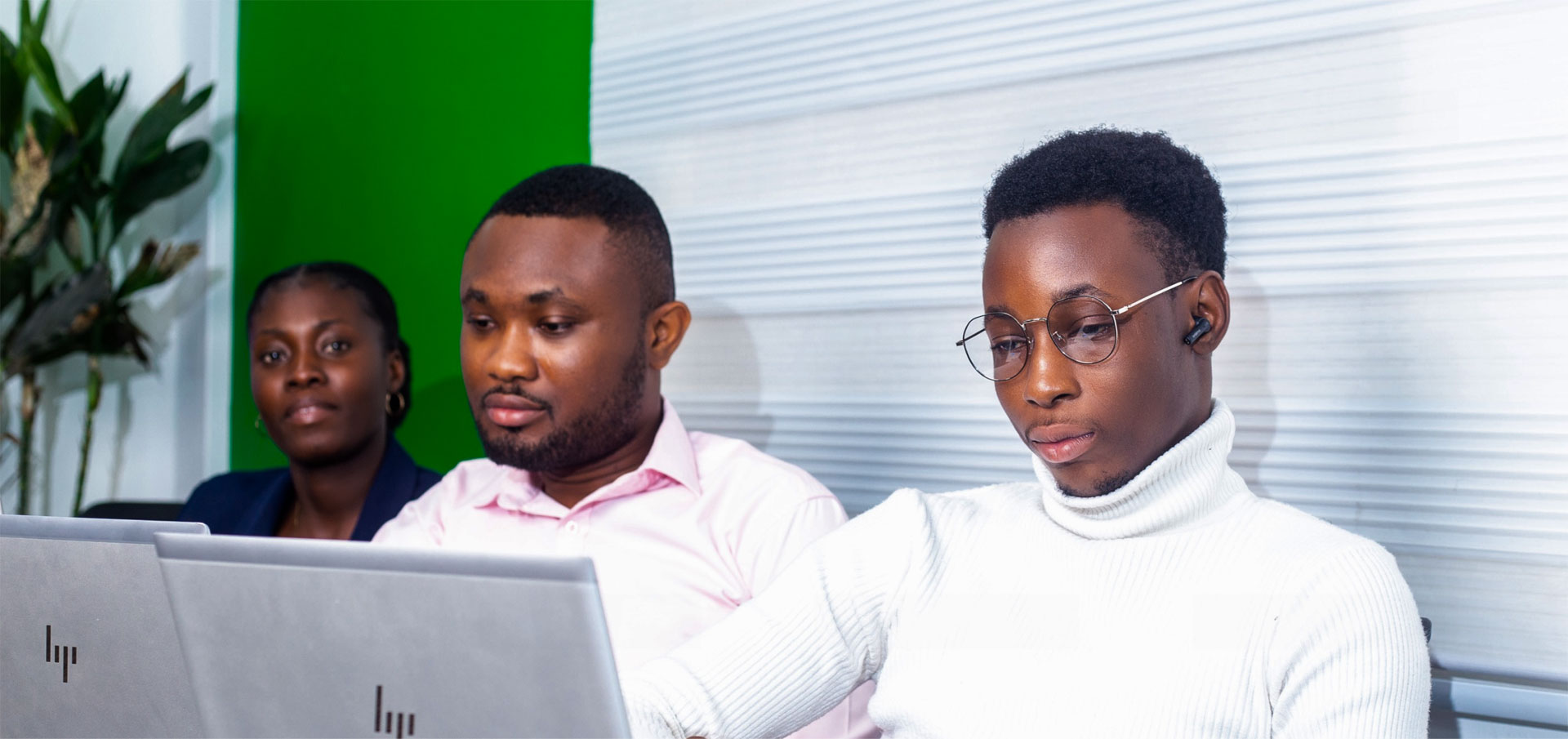 Three individuals are sitting in front of laptops. They are in a workspace, with a green panel on the left and horizontal blinds covering a window on the right.